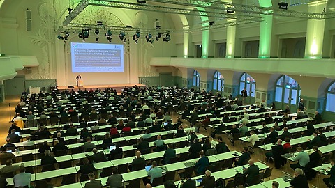 Ein großer Hörsaal mit vielen langen Tischreihen ist mit zahlreichen Personen besetzt, die einer Präsentation auf einer Bühne mit projizierter Folie folgen; der Saal ist hoch, weiß gestaltet und grün beleuchtet.
