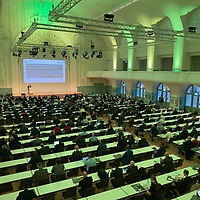 Ein großer Hörsaal mit vielen langen Tischreihen ist mit zahlreichen Personen besetzt, die einer Präsentation auf einer Bühne mit projizierter Folie folgen; der Saal ist hoch, weiß gestaltet und grün beleuchtet.