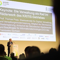 Ein Mann spricht auf einer Bühne am Rednerpult der Messe protekt vor einem großen Bildschirm, auf dem die Keynote von Prof. Thomas Popp mit Titel und Funktionstext angezeigt wird. Ein Mann spricht auf einer Bühne am Rednerpult der Messe protekt vor einem großen Bildschirm, auf dem die Keynote von Prof. Thomas Popp mit Titel und Funktionstext angezeigt wird.