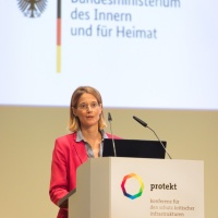 Eine Frau steht auf der protekt 2024 am Rednerpult mit Logo und Claim der Konferenz, im Hintergrund ist das Logo des Bundesministeriums des Innern und für Heimat projiziert. Eine Frau steht auf der protekt 2024 am Rednerpult mit Logo und Claim der Konferenz, im Hintergrund ist das Logo des Bundesministeriums des Innern und für Heimat projiziert.