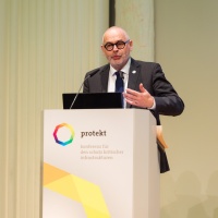 Ein Mann spricht auf der protekt 2024 mit Mikrofon und Presenter in der Hand am Rednerpult mit Konferenzlogo Ein Mann spricht auf der protekt 2024 mit Mikrofon und Presenter in der Hand am Rednerpult mit Konferenzlogo