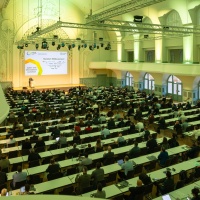 Ein großer Vortragssaal mit grüner Beleuchtung ist dicht mit Teilnehmenden besetzt, die einer Präsentation auf der Bühne folgen; auf der Leinwand ist das Logo der Messe protekt sichtbar. Ein großer Vortragssaal mit grüner Beleuchtung ist dicht mit Teilnehmenden besetzt, die einer Präsentation auf der Bühne folgen; auf der Leinwand ist das Logo der Messe protekt sichtbar.