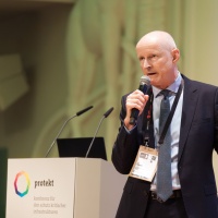 Ein Mann spricht auf der protekt 2024 mit Mikrofon und Presenter in der Hand am Rednerpult mit Konferenzlogo Ein Mann spricht auf der protekt 2024 mit Mikrofon und Presenter in der Hand am Rednerpult mit Konferenzlogo